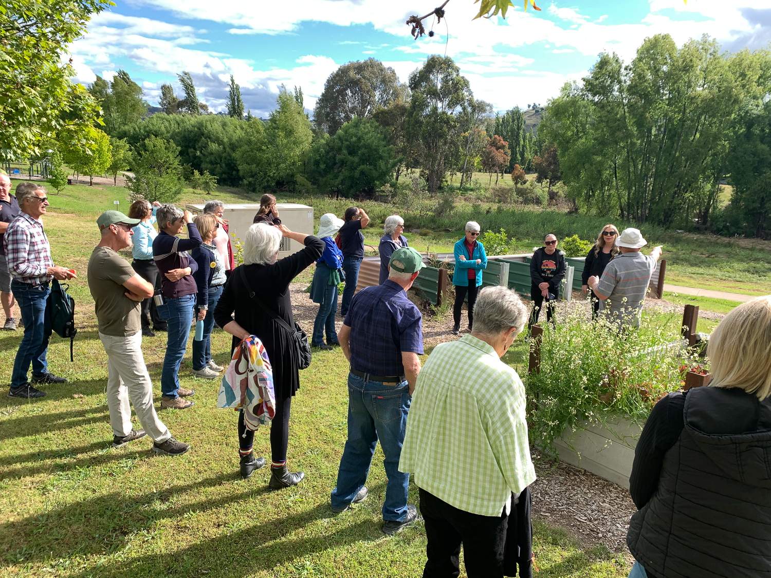 A group of people standing in sunshine in a community garden and looking towards the nearby creek.